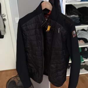Svart quiltad jacka Parajumpers - Svart quiltad jacka från Parajumpers med snygga detaljer som gul rem vid halsen och patch på ärmen. Jackan har dragkedja framtill, bröstficka med dragkedja och sportig look. Perfekt för dig som vill ha en stilren och funktionell jacka.Ända defekten är att dragkedjan är sönder