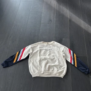 Randig sweatshirt från Kaotiko - Snygg sweatshirt från Kaotiko i storlek M. Tröjan är cremevit med långa ärmar i marinblått, dekorerade med röda, ljusblå och gula ränder. Rund hals och ribbade muddar. Perfekt för dig som gillar streetwear och färgglada detaljer.