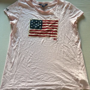 Ljusrosa t-shirt Polo Ralph Lauren XL - Ljusrosa t-shirt från Polo Ralph Lauren i bomull. Framsidan har en stor broderad amerikansk flagga med RL-broderi och ärmen har POLO-broderi. Klassisk rund hals och korta ärmar. Perfekt för en avslappnad och cool stil.