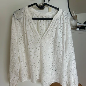 Vit broderad blus med v-ringning - Superfin vit blus från H&M med v-ringning och långa ärmar. Blusen har broderade detaljer och ett genombrutet mönster med blommor och cirklar. Tillverkad i mjuk bomull som känns luftig och fräsch. 