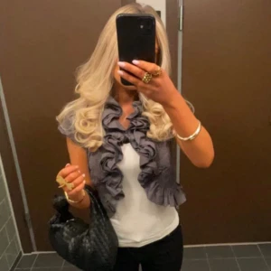 Grå bolero från Noa Noa - Supersnygg grå bolero från Noa Noa med korta ärmar och volangdetaljer längs krage, framkant och nederkant. Boleron har smockade partier vid midjan och är tillverkad i ett lätt bomullstyg. Perfekt för att lyfta en enkel outfit med extra edge. Sista tre bilderna är lånade. 