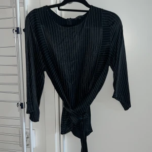 Marinblå randig blus med knyt - Snygg marinblå blus med tunna vita ränder och knyt framtill. Modellen har trekvartsärm och rund halsringning. Perfekt för dig som gillar stilrena plagg med en twist.