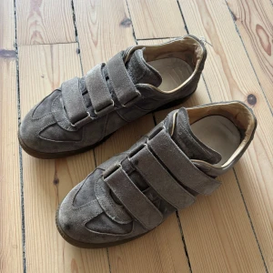 Margiela - Grå sneakers från Maison Margiela i mjuk mocka med tre breda kardborreband över foten. Skorna har en klassisk rund tå, gummisula med mönster undertill och beige innerfoder. Perfekt för dig som gillar stilren och unik design.
