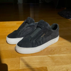 Axel arigato - Svarta sneakers från Axel Arigato i mocka med vit platt sula och diskret guldtext på sidan. Snygg minimalistisk design utan snörning, elastiskt band över foten och rund tå. Perfekt för dig som gillar stilrena och moderna skor.