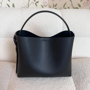 Svart bucket bag från Arket - Stilren svart bucket bag från Arket i slätt skinn. Väskan har en minimalistisk design med rundad form, bredt handtag och snyggt spänne på sidan. Perfekt för dig som gillar enkel och modern stil.