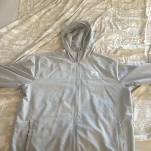 Ljusgrå vindjacka från Under Armour - Snygg ljusgrå vindjacka från Under Armour med huva och dragkedja hela vägen. Jackan har två sidofickor och meshfoder på insidan för extra komfort. Perfekt för dig som gillar sportig stil och vill ha något lätt och smidigt till vardagen.