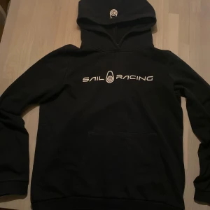 Svart Sail Racing hoodie 170 - Svart sail racing hoodie med känguruficka på magen och märke på huvan. Storlek 170. Bra skick, sparsamt använd, inga slitningar elr slitage. Nypris. Väldigt skönt material. Hör av dig vid frågor elr liknande🙌