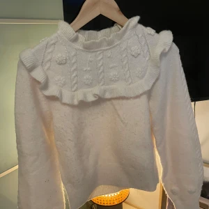Vit stickad tröja med volang & Other Stories - Supersöt vit stickad tröja från & Other Stories Paris Atelier. Tröjan har volangdetalj runt axlarna, blommönster och kabelstickning upptill samt långa ärmar. 