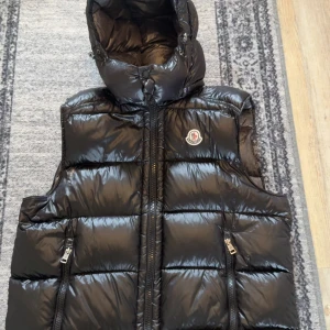 Svart dunväst från Moncler - Svart, glansig dunväst från Moncler med huva och dragkedja framtill. Västen har två fickor med dragkedja och klassisk Moncler-logga på bröstet. Tyvärr finns det ett litet hål men går att laga lätt och cartonen har lossnat i sömmen men sitter kvar bra ändå    går att laga. Går att ta av luvan. Stl 3, NFC tagg fungerar bild kan skickas  tyvärr inget kvitto, billigare vid snabb affär 