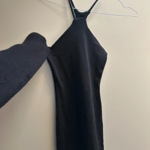 Svart tight klänning med smala band - En svart, tight klänning med smala axelband och hög halsringning. Klänningen är ärmlös och har en minimalistisk, stilren look som passar perfekt för en trendig och enkel outfit.