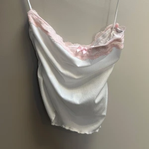 Vit ribbad linnetopp med spets - Supersöt vit ribbad linnetopp med tunna axelband, ljusrosa spetskant och en liten rosett framtill. Perfekt för en romantisk och feminin look. Mjuk och stretchig känsla.