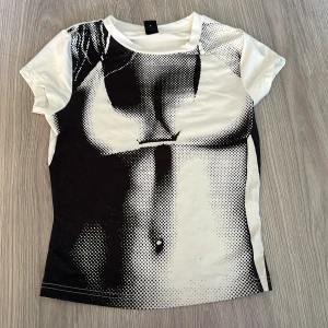 Vit t-shirt med svart grafiskt tryck - Cool vit t-shirt med ett stort svart grafiskt tryck framtill som föreställer en kropp i pop art-stil. T-shirten är kortärmad och har en klassisk rund halsringning. Materialet känns mjukt och stretchigt, perfekt för en avslappnad look.