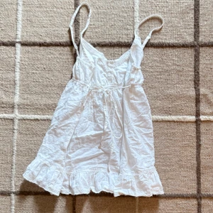 Vit lång babydoll linne med volang  - Söt vit linne med tunna axelband och volangkant nedtill. Linnen har knyt i midjan och spetsdetaljer vid halsringningen. Babydoll aktig. 