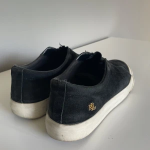Svarta sneakers från Ralph Lauren - Svarta sneakers från Ralph Lauren med logga i guld på sidan. Riktigt snygga och sköna! Lite slitna bak i hälen men märks inte när man har på dig dom💕💕