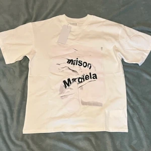 Maison Margiela vit t-shirt med tryck - Säljer en vit t-shirt från Maison Margiela med ett stort, abstrakt tryck och svart text på framsidan. T-shirten har klassisk passform och korta ärmar. Materialet är mjuk bomull och designen är riktigt edgy med den krossade logotypen framtill.