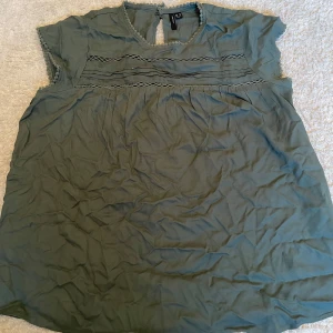 Grön blus från Vero Moda  - Snygg grön blus från Vero Moda i storlek M. Blusen har korta ärmar, rund halsringning med dekorativ kant och fina spetsdetaljer framtill. Lätt och luftig modell som knäpps i nacken med en liten knapp. Perfekt för en chill och stilren look.