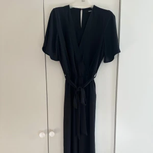 Svart jumpsuit från Bik Bok - Snygg svart jumpsuit från Bik Bok med omlott-design och v-ringning. Jumpsuiten har korta, fladdriga ärmar och ett knytband i midjan som ger en schysst siluett. Perfekt för dig som gillar stilrena och enkla plagg med lite extra edge