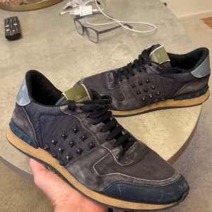 Valentino Rockrunner - Snygga svarta Valentino Garavani Rockrunner sneakers med ikoniska nitar på sidorna. Skorna har en mix av mocka och textil, svarta snören och kontrasterande beige sula. Perfekt för dig som vill sticka ut med en lyxig streetstyle.