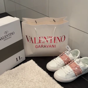Valentino Rockstud sneakers vit/rosa - Snygga Valentino Rockstud sneakers i vitt skinn med breda ljusrosa band och ikoniska nitar. Skorna har rund tå, platt sula och coola detaljer med pyramidnitar både på sidan och bak. Perfekta för dig som vill sticka ut med exklusiv streetstyle.