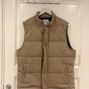 Beige quiltad dunväst från Zara - Snygg beige dunväst från Zara med quiltad design och hög krage. Västen har två fickor framtill och stängs med tryckknappar. Perfekt att slänga över en hoodie eller tröja för en chill look.