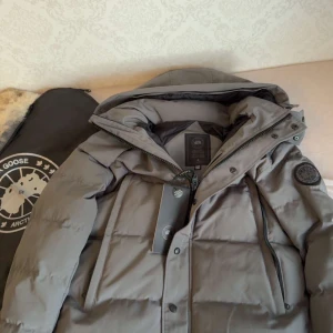 Grå Canada goose wyndham black label - Säljer en grå dunjacka från Canada Goose med klassisk patch på ärmen. Jackan har huva, dragkedja och knappar framtill samt två stora fickor. Perfekt för kalla dagar och riktigt snygg till vinterlooken. Päls ingår och det är storlek M