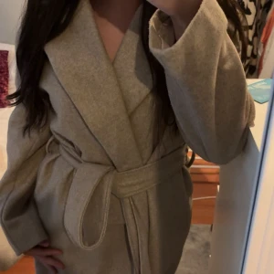 Beige kappa med bälte och omlott - Snygg beige kappa med bred krage och omlottmodell. Jackan har ett matchande bälte i midjan som knyts för en clean och stilren look. Perfekt för dig som gillar klassiska vibes och vill hålla dig varm under kyliga dagar.
