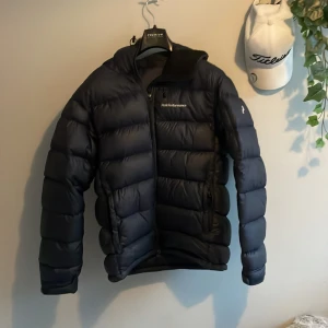 Mörkblå Peak Performance dunjacka S - Säljer en mörk blå  dunjacka från Peak Performance i storlek S. Jackan har en puffig design med horisontella sömmar, huva och dragkedja framtill. Perfekt för kalla dagar och riktigt snygg till streetwear-stilen.