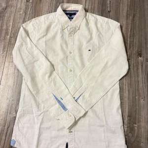 Vit slim fit skjorta Tommy Hilfiger - Vit skjorta från Tommy Hilfiger i slim fit-modell med stretch. Klassisk krage, knappar framtill och diskret logga på bröstet. Snygga blå detaljer på insidan av kragen och manschetterna. Perfekt för en clean och stilren look.