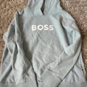 Ljusblå hoodie från BOSS - Snygg ljusblå hoodie från BOSS med stor vit logga framtill. Tröjan har huva och långa ärmar, tillverkad i mjuk bomull. Perfekt för en chill och stilren look.