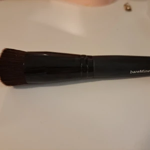 bareMinerals foundationborste - Snygg foundationborste från bareMinerals med svart handtag och täta, mjuka borststrån i mörkbrunt. Borsten har en platt och rundad form som gör det enkelt att applicera och buffa in foundation för ett jämnt resultat. Perfekt för dig som vill ha en proffsig finish.