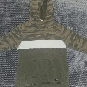 Camouflage hoodie från KappAhl - Säljer en grön hoodie från KappAhl med camouflagemönster på huva och överdel, vit bred rand över bröstet och nedre delen i olivgrönt. Tröjan har magficka och långa ärmar, perfekt för chill dagar. Materialet är mjuk bomull och polyesterblandning.