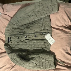 Quiltad grön jacka från Zara - Snygg quiltad jacka från Zara i en dämpad grön nyans. Jackan är croppad med långa ärmar, dragkedja och knappar framtill. Perfekt för lager-på-lager och har en modern, avslappnad vibe. Passar dig som gillar trendiga och bekväma plagg.