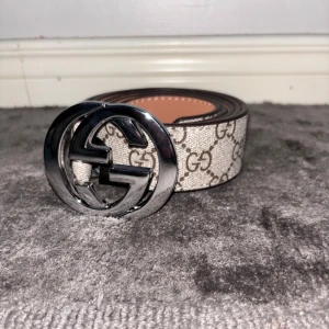 Gucci GG beige bälte med silverdetalj - Snyggt bälte från Gucci i beige med klassiskt GG-mönster och stor, rund spänne i silver. Perfekt accessoar för att lyfta vilken outfit som helst med en lyxig touch. Bältet har en stilren design och är tillverkat i skinn med canvasdetaljer.
