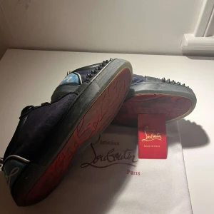 Christian Louboutin svarta sneakers med nitar - Säljer ett par svarta Christian Louboutin sneakers med ikoniska röda sulor och coola svarta nitar på tån. Skorna har snörning, detaljer i blått och vitt på hälen och är tillverkade i mocka och skinn. Perfekt för dig som vill sticka ut med lyxig streetstyle.