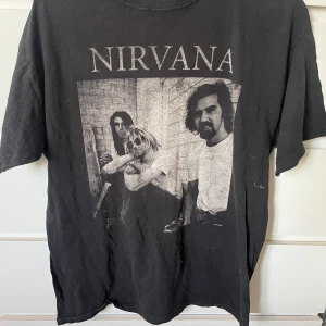 Svart Nirvana t-shirt med tryck - Cool svart t-shirt med stort Nirvana-tryck på bröstet och ikoniskt svartvitt foto av bandet. Klassisk passform med rund hals och korta ärmar. Perfekt för dig som älskar grunge och vill ha en statement-tee i garderoben.