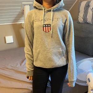Grå hoodie från GANT med tryck - Säljer en grå hoodie från GANT i storlek S. Använts fåtal gånger förut. Tröjan har huva med vita snören, magficka och ett broderat GANT-tryck med amerikansk sköld på bröstet. Mjuk bomullsmix och ribbade muddar vid ärmslut och nederkant. Perfekt för chill dagar.