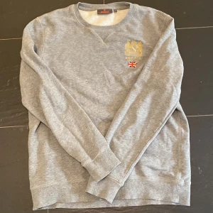 Grå sweatshirt från Morris med brodyr - Grå sweatshirt från Morris med guldfärgad brodyr och brittisk flagga på bröstet. Klassisk rund halsringning och långa ärmar. Mjuk och skön bomullsblandning, perfekt för chill dagar eller när du vill ha en clean look.