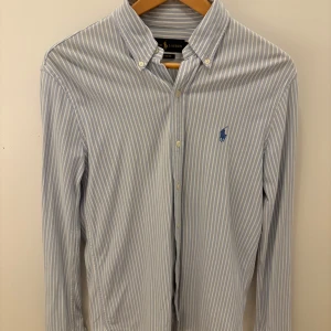 Randig skjorta från Ralph Lauren - Klassisk blå- och vitrandig skjorta från Ralph Lauren med broderad logga på bröstet. Skjortan har lång ärm, knappar framtill och button-down krage. Tillverkad i mjuk bomull och passar perfekt till en clean och stilren look.