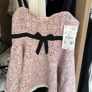 Rosa tweed korsettopp med rosett - Supersöt korsettopp i rosa och svart tweed med svarta axelband och en  svart rosett framtill. Toppen har en lätt utsvängd form och dragkedja bak🩷 