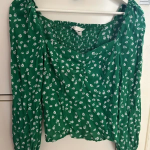Grön blommig blus med puffärm - Snygg grön blus med vita små blommor och puffiga långärmar. Smockad upptill för en skön passform och fyrkantig halsringning som ger en trendig vibe. Perfekt att styla med jeans eller kjol för en cool look.