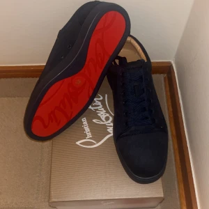 Christian Louboutin svarta sneakers - Snygga svarta sneakers från Christian Louboutin i mocka med klassisk röd sula och broderad logga på hälen. Skorna har rund tå, platt sula och svarta skosnören. Perfekt för dig som vill sticka ut med exklusiva detaljer och lyxig känsla.