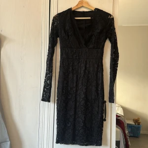 Svart spetsklänning från NLY One - Supersnygg svart spetsklänning från NLY One i storlek S. Klänningen har långärmade ärmar i transparent spets, v-ringad omlottfront och figurnära passform. Den är knälång och har ett elegant spetsmönster över hela klänningen. Perfekt för dig som vill ha en chic look.