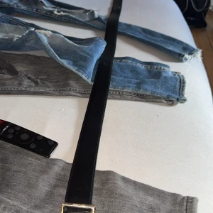 Svart skärp från Amiri med gulddetalj - Säljer ett svart bälte från Amiri med en fet guldfärgad logotyp som spänne. Bältet är stilrent och har en slät, klassisk form i äkta skinn. Kommer med originalbox och kvitto. Perfekt för dig som vill ha en lyxig vibe och visa upp märket.