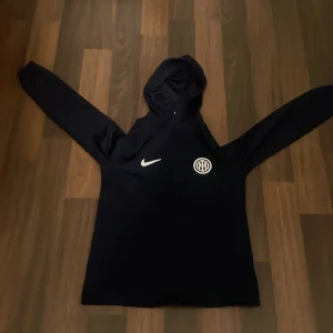 Mörkblå Nike Inter hoodie Dri-FIT - Säljer en mörkblå hoodie från Nike med Inter-logga på bröstet och Dri-FIT material som andas. Tröjan har huva, långa ärmar och en clean look med vit Nike-swoosh. Perfekt för dig som gillar fotboll och vill ha en sportig stil.