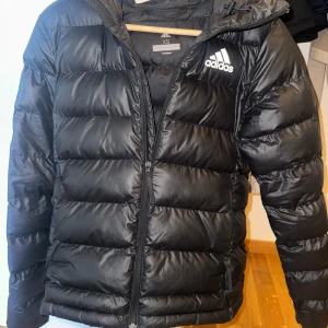 Svart pufferjacka från Adidas XS - Svart pufferjacka från Adidas i storlek XS. Jackan har huva, dragkedja framtill och Adidas-logga på bröstet. Tillverkad i glansig polyester med horisontella quiltade sektioner. Perfekt för kalla dagar och har en sportig vibe. Jackan är i riktig bra skick inga skador eller så men den började läcka dun vid ärmen förut som vi sydde och det syns knappt.