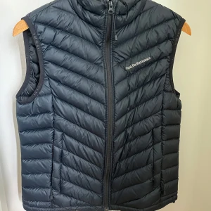 Svart dunväst Peak Performance - Snygg svart dunväst från Peak Performance med quiltad design och dragkedja framtill. Västen har hög krage och diskret logga på bröstet. Perfekt att slänga över en hoodie eller tröja när det är kyligt ute. Västen är i storlek M