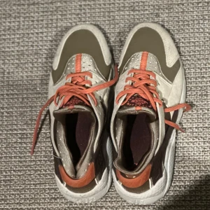 Nike Huarache sneakers - Nike sneakers i beige, brunt och olivgrönt med orange detaljer och snörning. Ovandel i mocka och syntet, vit sula och cool retrodesign. Perfekta för dig som vill sticka ut med snygga färgkombos och sportig vibe.