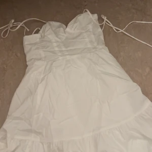 Vit kort klänning med knytband - Supersöt vit kortklänning med smala axelband som knyts. Klänningen har en söt hjärtformad urringning och volangig kjol. Perfekt för sommarens varma dagar och har en luftig känsla.