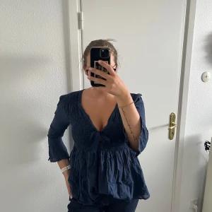 Blå Odd Molly blus med broderi - Supersöt blå blus från Odd Molly med v-ringning och knyt framtill. Blusen har smockad överdel, volang nedtill och trekvartsärmar. Snyggt broderat fjärilsmärke på ena ärmen och tunna ränder i tyget. 