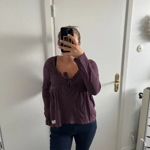 Lila Odd Molly blus med knyt och smock - Supersöt plommonfärgsd blus från Odd Molly med smockad överdel och knyt framtill. Långärmad och tillverkad i mjuk bomull. Söt broderad detalj i nacken och lätt volang vid halsringningen. Perfekt för dig som gillar unika detaljer och skön passform.